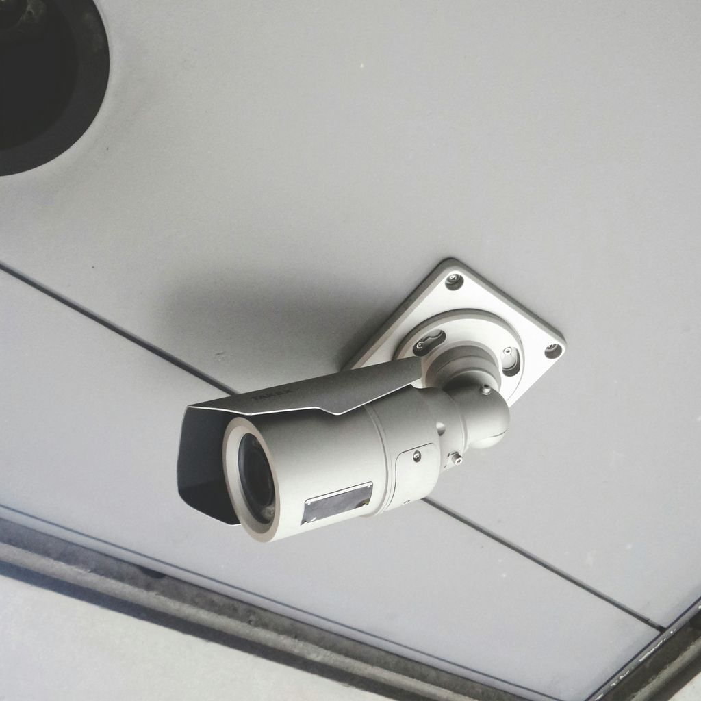 The image shows a security camera mounted on a ceiling. It's a bullet-style CCTV camera, fixed to a metal bracket with screws. The camera appears to be positioned to monitor an indoor or sheltered area, likely for surveillance purposes.