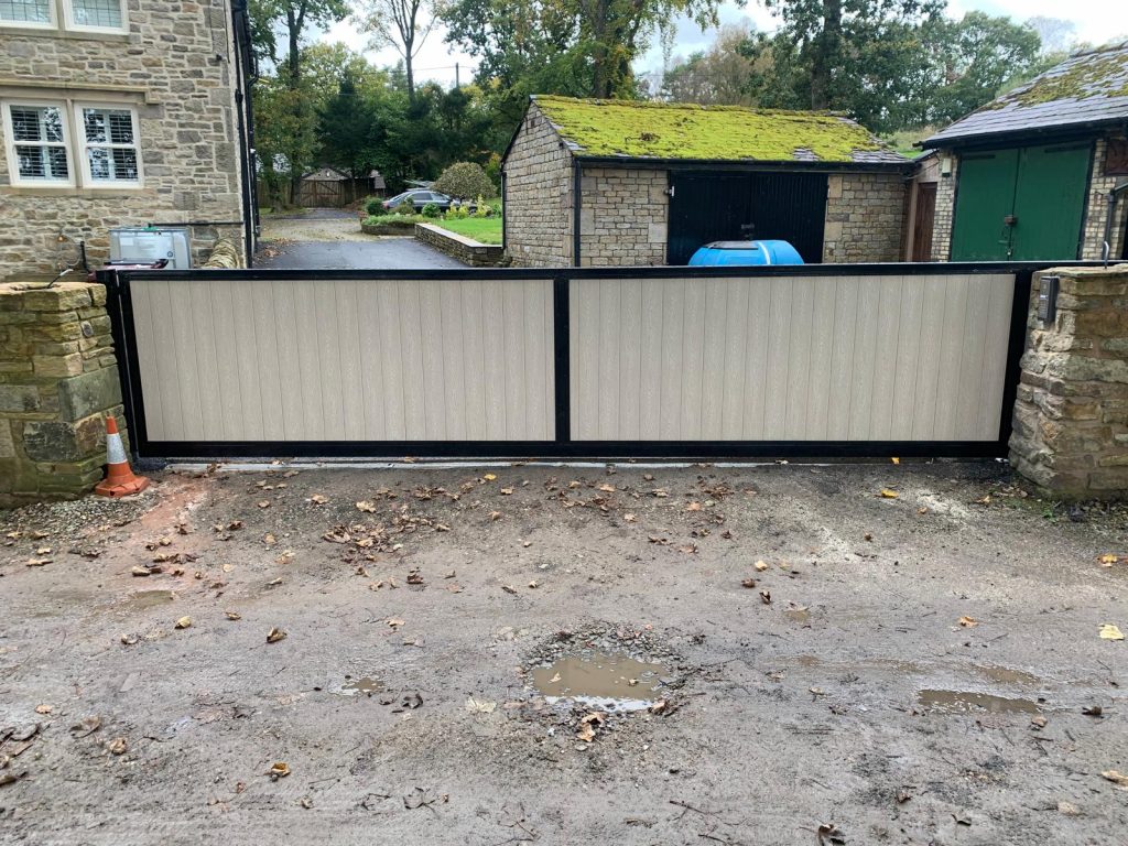 A wide, modern sliding driveway gate with a black metal frame and light-coloured vertical panel inserts, installed between two stone pillars at the entrance of a rural property with stone buildings in the background.