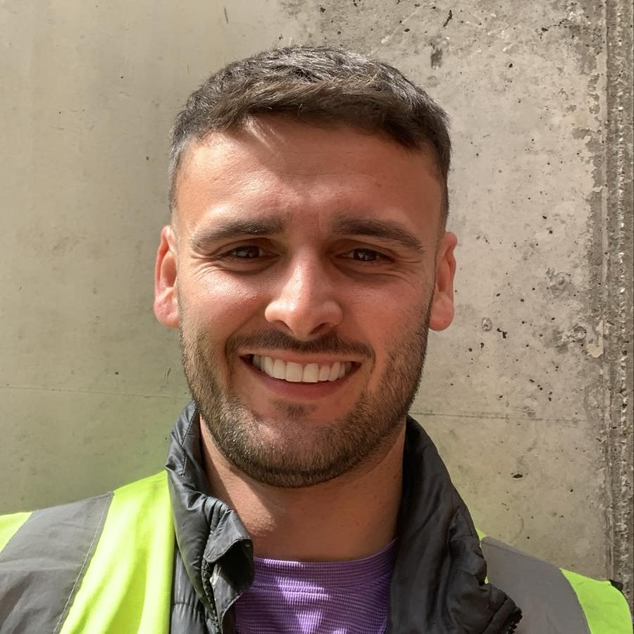 Sam Milton, part of our team, pictured on-site in a high-visibility vest, smiling in front of a concrete wall.