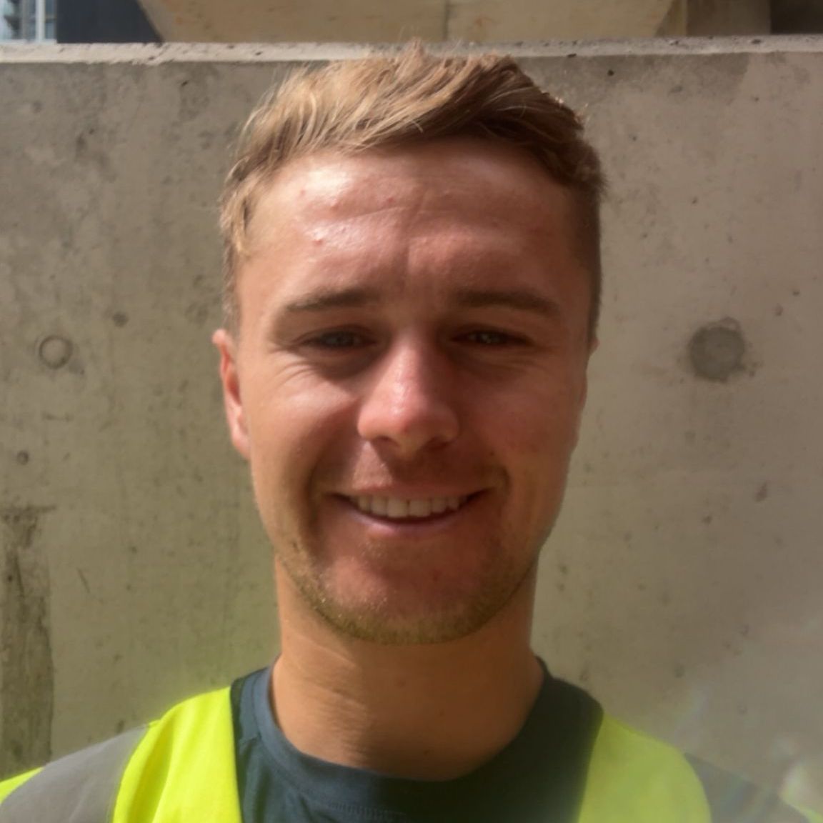 Sam Rigby, part of our team, pictured on-site wearing a high-visibility vest and smiling in front of a concrete wall.