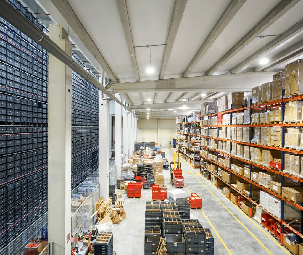 Large industrial warehouse interior with high shelves stacked with boxes and containers, wide aisles, and various pallets and equipment on the floor.
